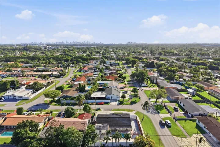 Aerial perspective of a suburban neighborhood featuring mature landscaping, paved street grids, residential lots, and a distant cityscape skyline