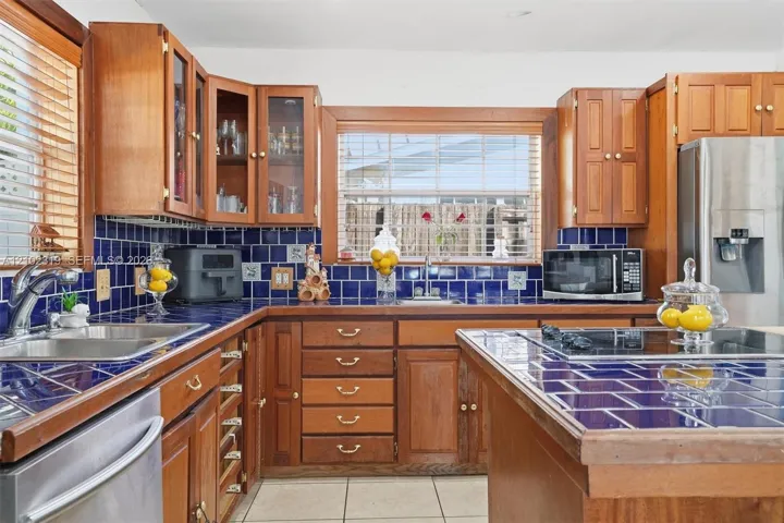 Kitchen featuring stained wood cabinetry, blue tiled countertops and backsplash, stainless steel double-basin sink, center island with cooktop, and stainless steel refrigerator