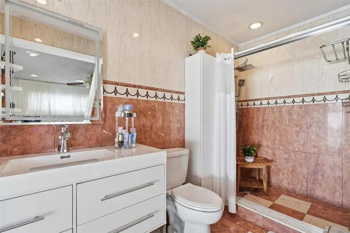 Bathroom featuring stone-tiled walls with decorative border inlay, a white vanity with integrated basin, polished metal hardware, a glass-framed mirror, and a walk-in shower area with stone flooring