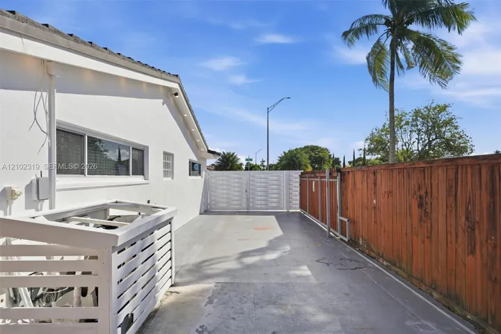 Exterior side yard featuring white stucco siding, a concrete paved surface, a white slatted privacy screen, and a tall wood-plank perimeter fence