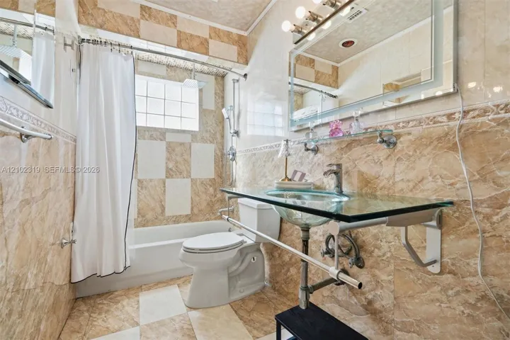 Bathroom featuring floor-to-ceiling tiled walls in a checkerboard pattern, glass vanity countertop with integrated basin, polished metal faucet, glass block window, and large rectangular mirror with an integrated light fixture