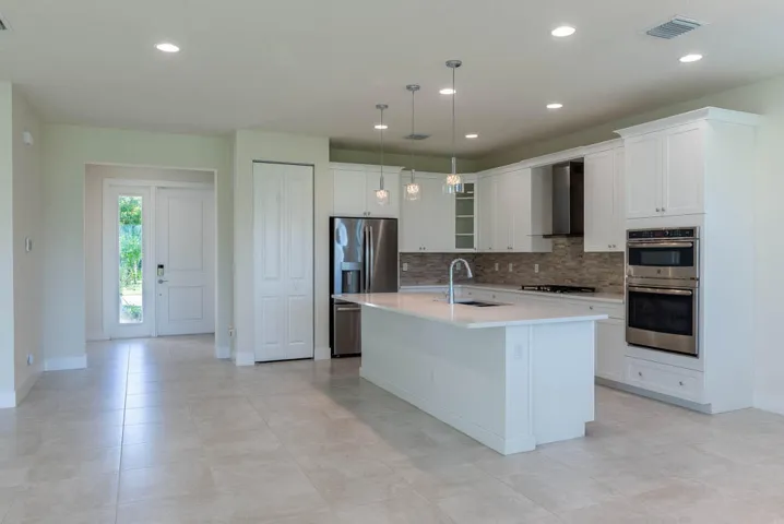Open-concept living area with light-toned tile flooring, recessed lighting, white shaker-style cabinetry, a central island with a sink, and stainless steel appliances