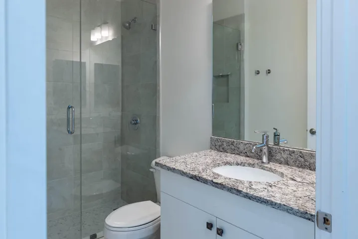 Modern bathroom featuring a frameless glass shower enclosure with tile surround, dual vanity with speckled stone countertop, chrome fixtures, and ample mirror lighting