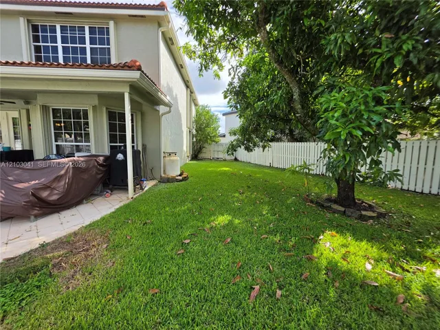 Expansive green space featuring a white picket fence, mature trees, and a covered patio with tiled flooring