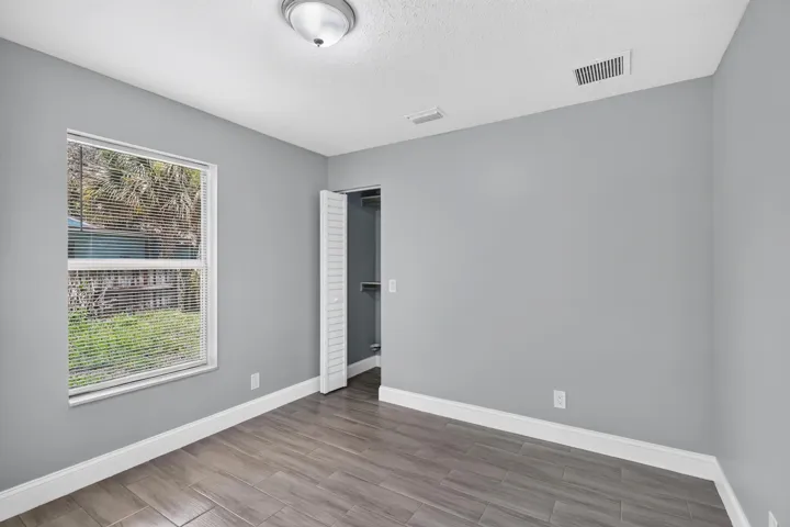 Spacious room featuring wood-finish tile flooring, a large window with blinds, light gray walls, a ceiling-mounted light fixture, and a closet with louvered bi-fold doors
