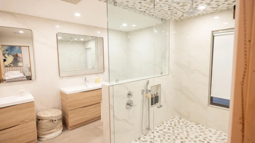 Spacious bathroom featuring two wood-finish vanities with rectangular sinks, a glass-enclosed shower with a rainfall showerhead, and hexagon-tiled shower floor and ceiling