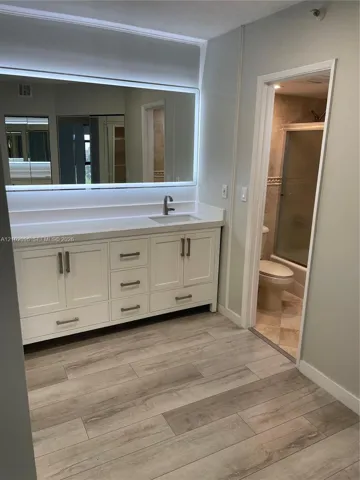 White vanity with brushed metal hardware, solid-surface countertop, rectangular undermount sink, and oversized wall-mounted mirror with integrated perimeter lighting, complemented by wood-finish flooring