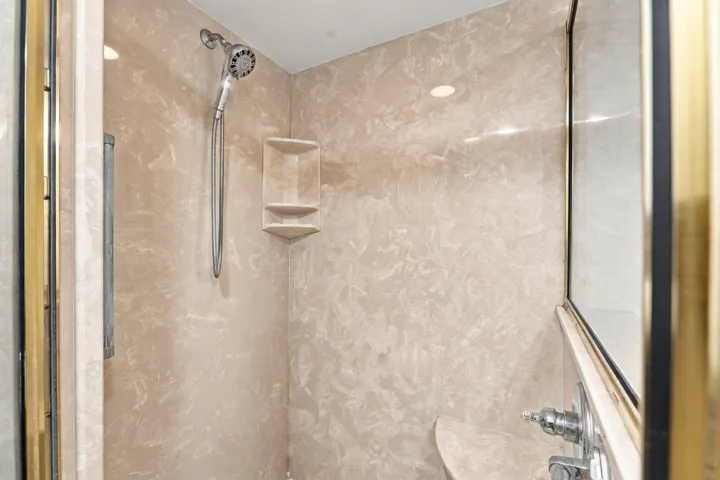 Spa-inspired shower enclosure featuring polished marble-finish tile walls, chrome rainfall showerhead with handheld spray fixture, recessed shelving for storage, and frameless glass enclosure with brushed metal hardware