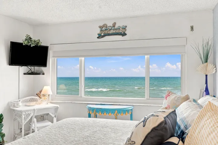 Oceanfront bedroom showcasing expansive floor-to-ceiling windows framing direct water vistas, white wicker furnishings, neutral bedding, and a mounted flat-screen television