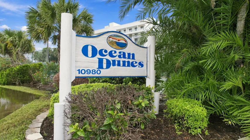 Signage featuring property name, lush landscaping, and mature palm trees