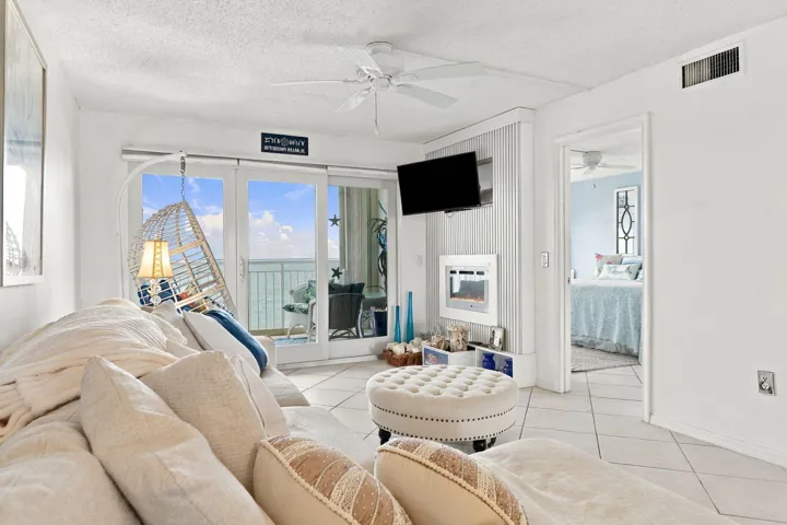 Bright, open living area featuring floor-to-ceiling glass doors and windows with ocean vistas, neutral tile flooring, ceiling fan, wall-mounted flat-screen television, and access to a furnished balcony overlooking the waterfront