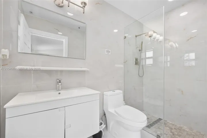 Bathroom featuring a glass-enclosed shower, wall-mounted shower system, white vanity with integrated sink, and white large-format wall tiles