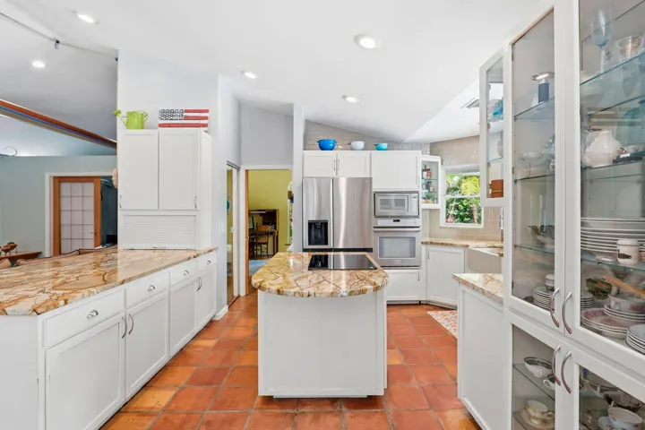 Kitchen featuring an island, white cabinetry, integrated stainless steel appliances, natural stone countertops, and terra cotta tile flooring