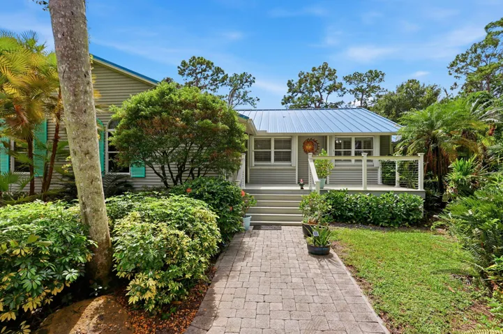 Stone paver walkway leading to a front porch with white railing, a light gray exterior, and a blue metal roof