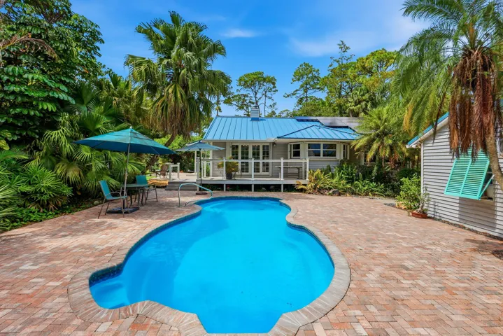 Freeform pool with brick paver surround, lush tropical landscaping, and a pale blue metal roof