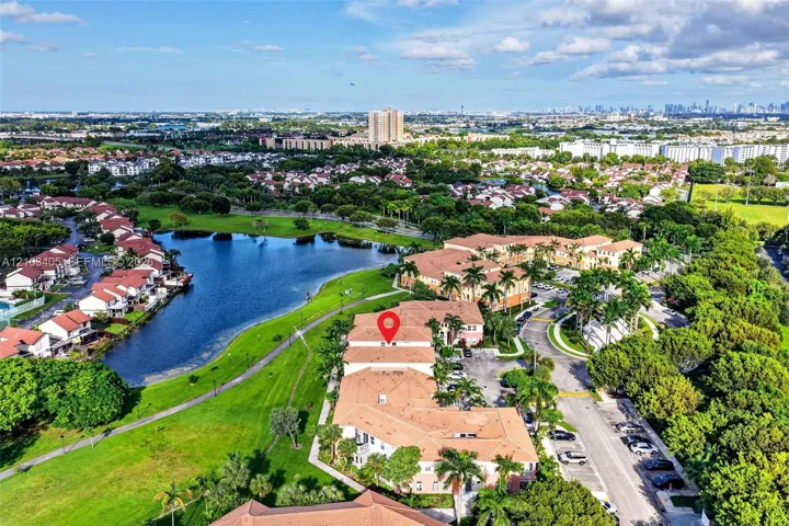 Aerial perspective featuring a multi-story building with a clay tile roof, situated near a lake, green space, and palm trees