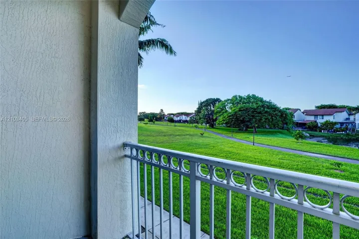 Covered balcony featuring textured stucco pillars and decorative metal railings overlooking a landscaped lawn with mature trees and a paved path