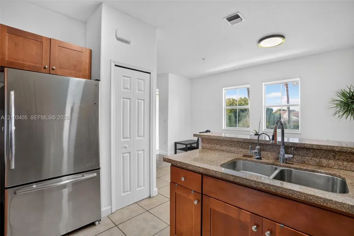 Kitchen featuring wood cabinetry, stone countertops, stainless steel dual-basin undermount sink with a gooseneck faucet, stainless steel refrigerator, and tile flooring