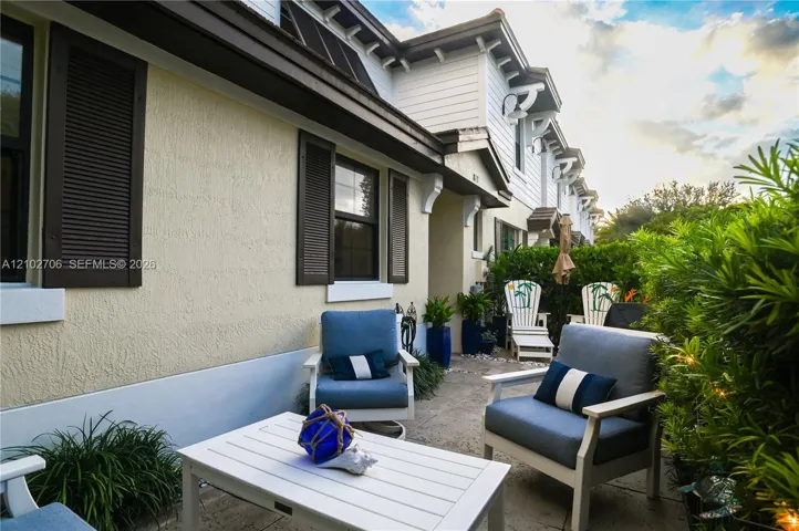 Exterior facade featuring light-colored stucco siding, dark-toned shutters, architectural roof detailing, and a stone-paved patio area bordered by mature shrubbery