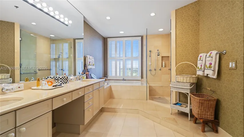 Extended vanity with dual integrated sinks and stone countertop, soaking tub encased in stone tile, walk-in glass-enclosed shower with multiple showerheads, and interior window shutters