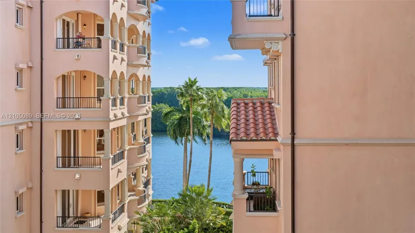 Stucco exterior with multi-level arched balconies, metal railings, column supports, and clay tile roof accents, featuring a water backdrop with palm trees