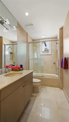 Bathroom featuring a light-toned vanity with an integrated basin, expansive wall-to-wall mirror, tiled flooring, and a glass-enclosed shower and tub combination with wall-mounted plumbing fixtures