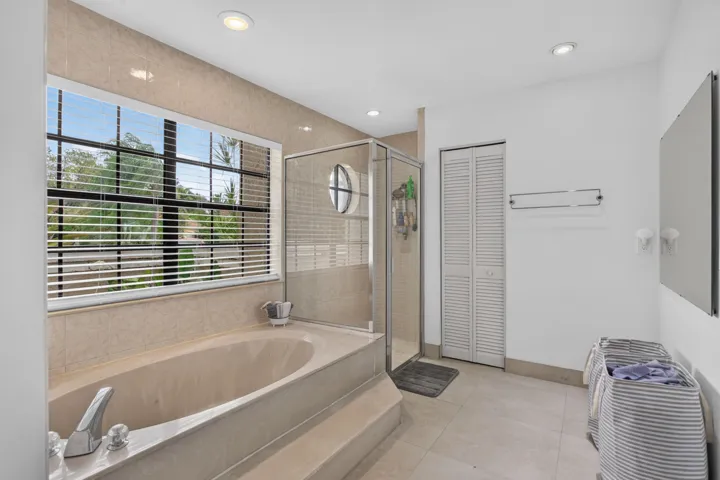 Recessed lighting, large window with horizontal blinds, built-in bathtub with tiled surround, glass-enclosed shower, and louvered bifold doors