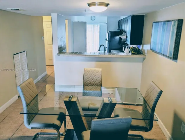 Dining area featuring neutral tile flooring and a pass-through breakfast bar with tiled countertop, opening to a kitchen equipped with dark-finish cabinetry