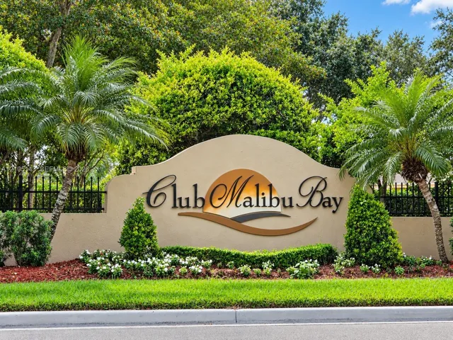 Club Malibu Bay entrance sign with a sculpted facade, wrought iron fencing, lush landscaping, and mature palm trees