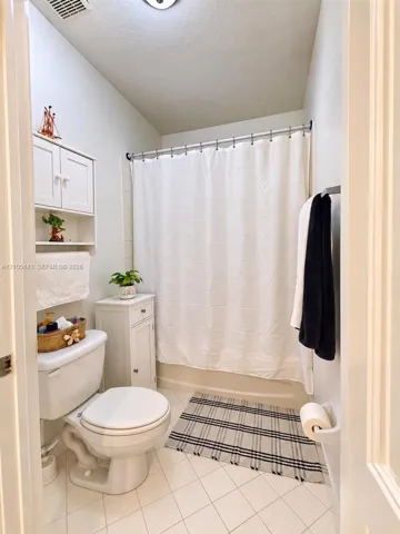 White tile flooring, a built-in toilet, a white wall-mounted storage cabinet, and a shower with a white curtain
