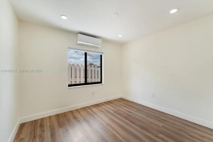 Recessed ceiling lighting, wood-finish flooring, single window, roller shade, and wall-mounted air conditioning unit