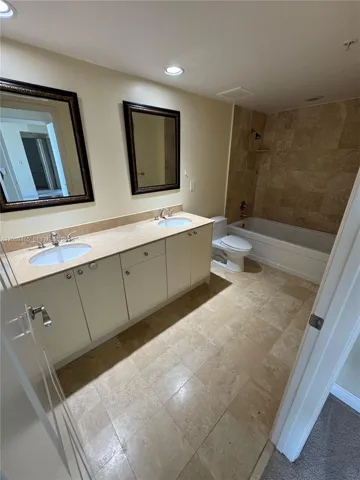 Dual-sink vanity with light-toned countertop, stone-tile flooring, and stone-tiled bathtub surround