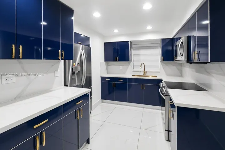 Contemporary kitchen featuring dark blue cabinetry with gold-tone hardware, white countertops, stainless steel appliances, and recessed lighting