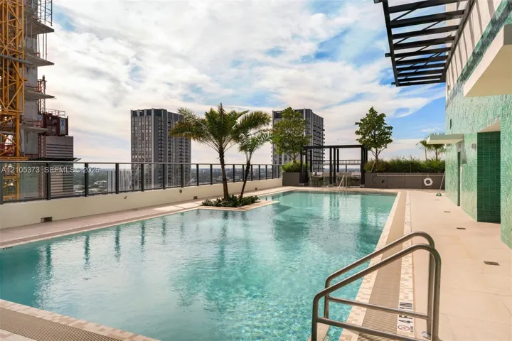 Rooftop pool with clear water, glass perimeter railing, surrounding cityscape, integrated palm trees, and textured green exterior wall