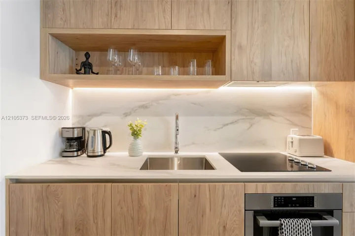 Light wood-finish cabinetry with an open shelf, undermount stainless steel sink, integrated cooktop, stainless steel built-in oven, and white marble-patterned backsplash