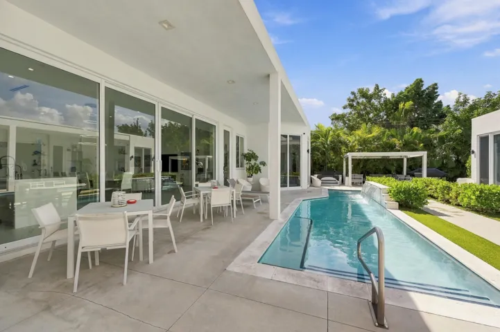 Expansive outdoor living area featuring a covered patio, a rectangular swimming pool with integrated steps, a white pergola, and manicured landscaping