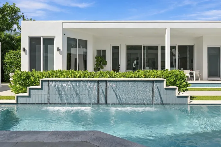 Contemporary exterior with a flat roofline, featuring an integrated pool with a waterfall element, expansive sliding glass doors, and a covered patio area