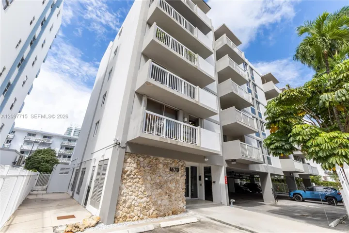 Multi-story building featuring balconies with white railings, a stone-clad lower facade, a covered parking area, and mature tropical landscaping