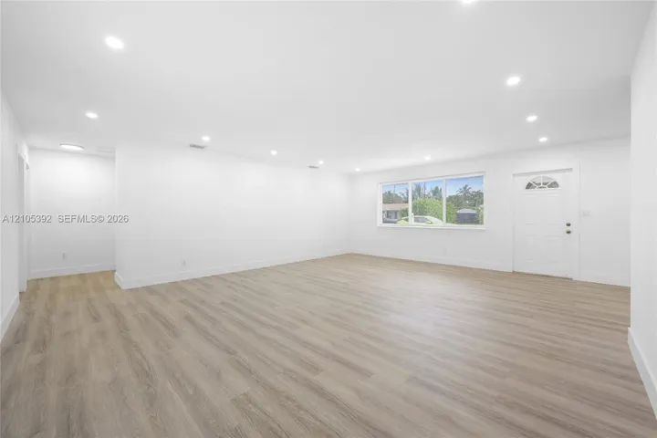 Expansive living area with wood-finish flooring, recessed lighting, large window, and white entry door with decorative glass insert