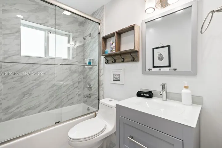 Built-in tub with glass enclosure, large format wall tile, single vanity with integrated sink, wall-mounted mirror, and wall-mounted light fixture