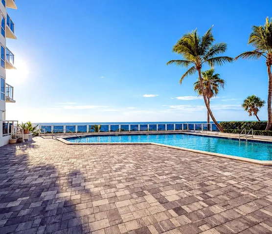 Resort-style pool with integrated steps, surrounded by an expansive paver deck, clear glass railing, and mature palm trees