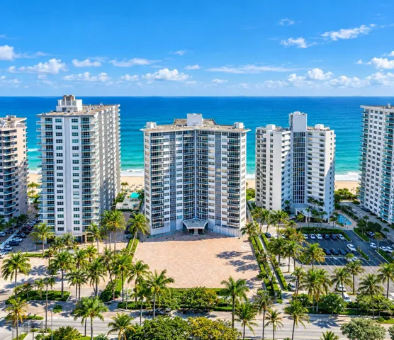 Oceanfront high-rise building with multiple balconies, extensive paved entry, and lush landscaping