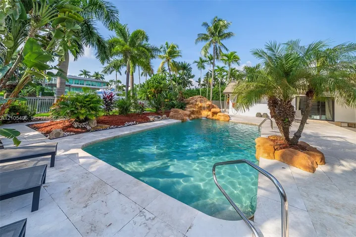 Resort-style pool with rock formation waterfall, surrounded by extensive patio pavers and lush tropical landscaping