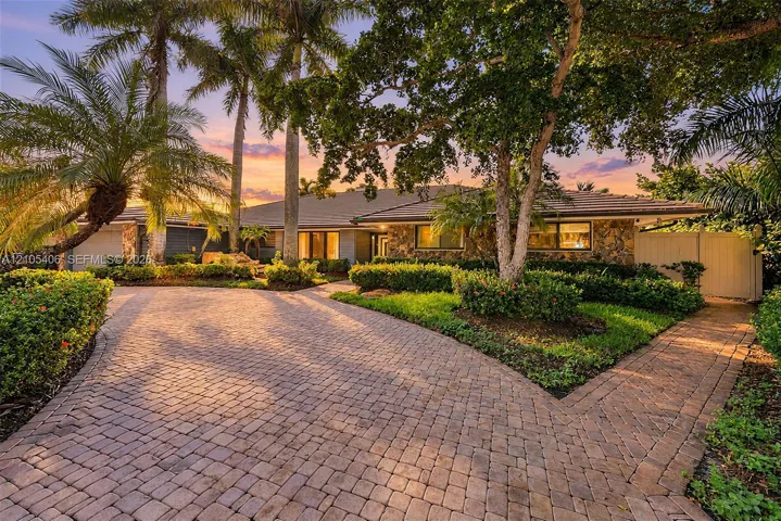 Expansive paver driveway with split path, lush landscaping, and mature palm trees framing a stone-clad exterior