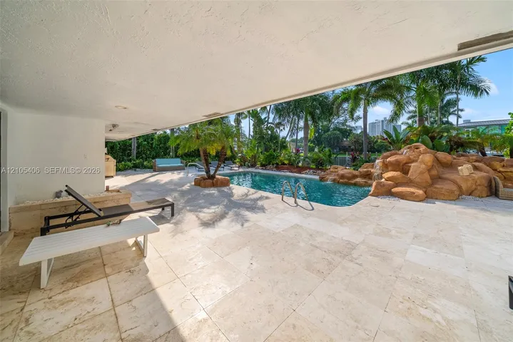 Expansive covered patio featuring light-toned tile flooring, directly accessing a custom pool with a rock waterfall feature and integrated steps