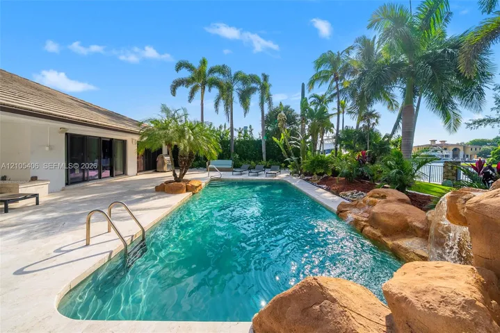 Rectangular swimming pool with rock waterfall feature, surrounding paver patio, and mature tropical landscaping