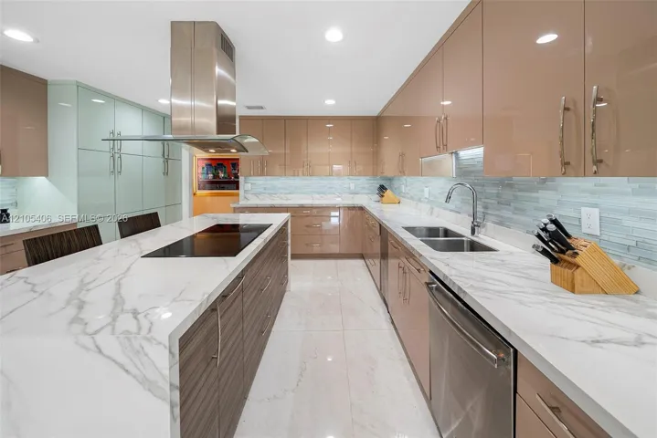 Kitchen featuring a large island with an integrated cooktop, white marble-style countertops, glossy tan cabinetry, a stainless steel range hood, and a dual basin sink