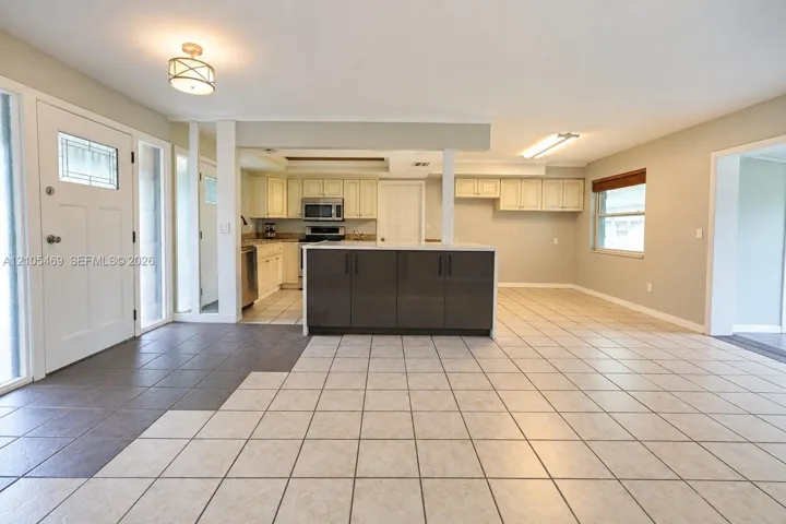 Open-concept living area with large format tile flooring, a kitchen island with dark cabinetry, stainless steel appliances, and a window with blinds