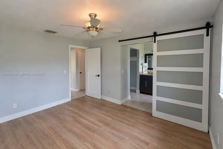Wood-finish flooring, ceiling fan with integrated lighting, a modern barn door with frosted glass panels, and a bathroom entrance featuring a dark vanity with a light-toned countertop