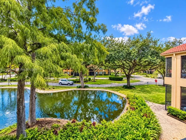 Waterfront property featuring a pond, mature landscaping, paved walkway, and multi-story building with a red-tiled roof and screened enclosures